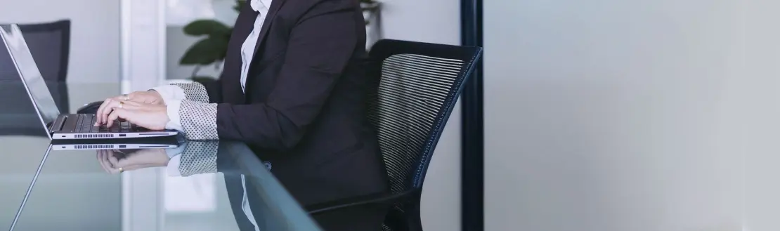 Person im Business-Anzug arbeitet an einem Laptop an einem Glastisch in einem modernen Büro.